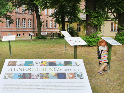 Ansicht der Open-Air-Ausstellung Auserlesenes. Acht in den Rasen eingelassene Stelen mit Schautafeln vor einem Backsteingebäude. Unter einer Stele stehendes Kind hält sich die Augen zu.