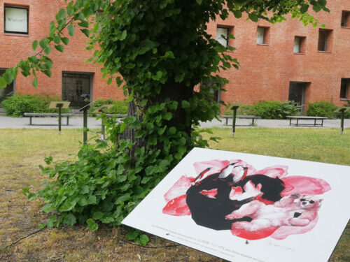 Ansicht der Open-Air-Ausstellung Auserlesenes. In den Rasen eingelassene Stelen mit Schautafeln vor einem Backsteingebäude. Im Vordergrund: Erzählbild mit Eisbär von Susanne Janssen.