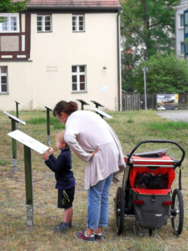 Ansicht der Open-Air-Ausstellung Auserlesenes vor dem Museum Köpenick. Mutter mit Kind nebst Fahrradanhänger betrachten eine in den Rasen eingelassene Schautafel auf Stele.