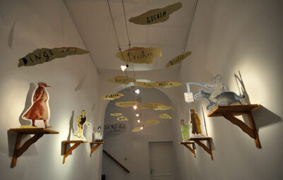 Foyergestaltung zur Ausstellung “Die Weite hinter den Worten”. Illustrations-Pappfiguren auf Wandkonsolen, Wörter hängen von der Decke.