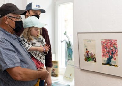Erwachsene und Kinder besuchen die Ausstellung “Von Anna Humpelhexe bis Zacharias Zappelbein” und betrachten Bilder von Ingeborg Friebel.