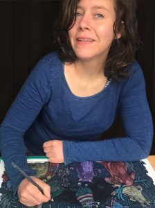 Illustratorin Iris Anemone Paul beim Gestalten ihres Erzählbildes. Sie trägt braune halblange Haare, einen blauen Pullover und hält einen Buntstift in der Hand.